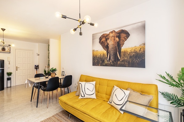 A living room with a yellow sofa, a dining table with chairs, a chandelier, and a large elephant painting on the wall.