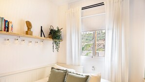 Ett sovrum med en säng, kuddar, en hylla med böcker och en växt, ett fönster med gardiner och utsikt över grönområden utanför.