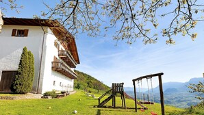 A white building with a wooden balcony, a swing set, and a clear view of the mountains.