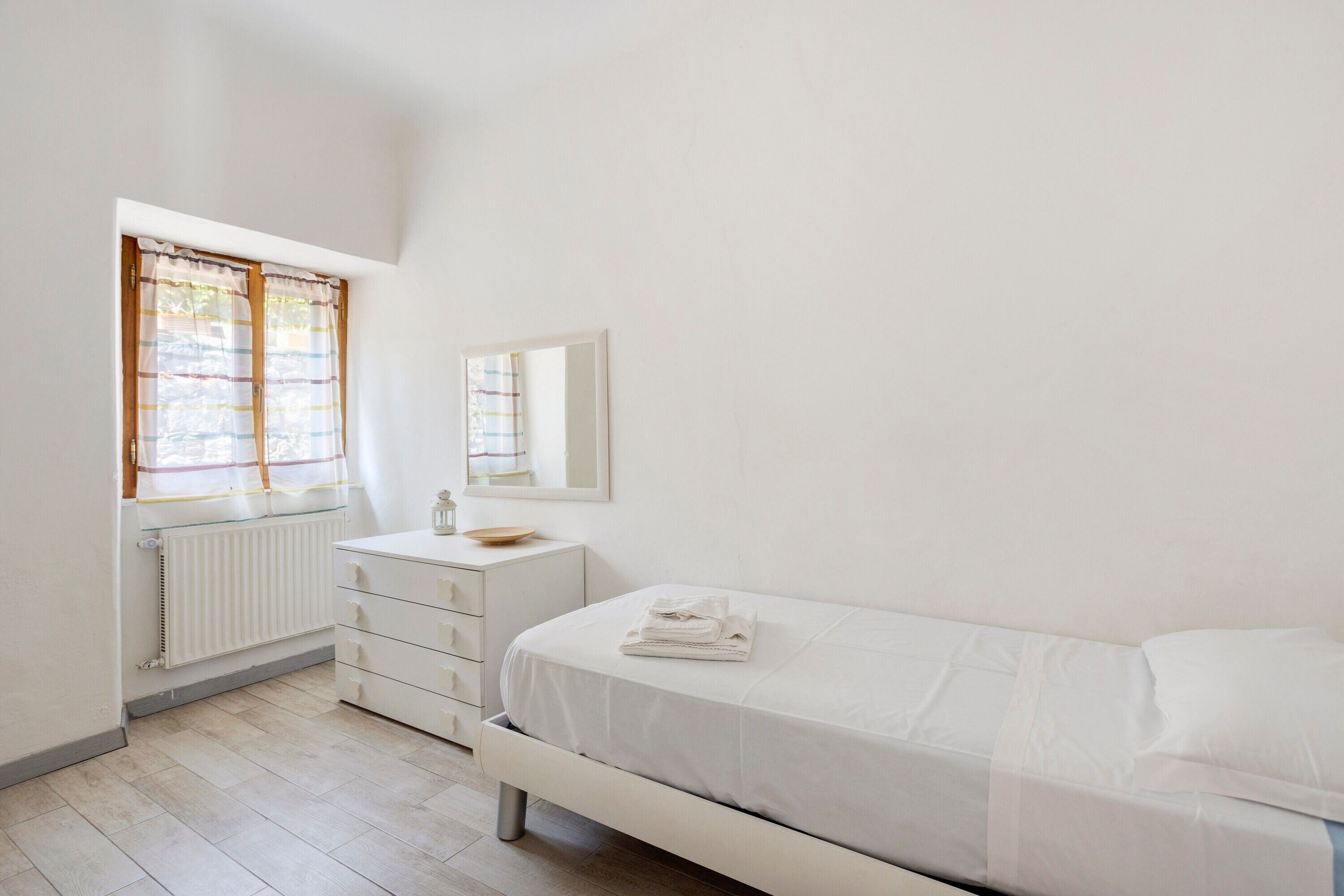 Une chambre à coucher comprenant un lit, une commode, un miroir, un radiateur et une fenêtre avec des rideaux.