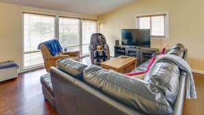 Een woonkamer met een leren bank, een fauteuil, een salontafel, een televisie en grote ramen met jaloezieën.