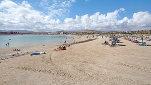 A beach with people sunbathing and swimming, umbrellas, and a clear sky.