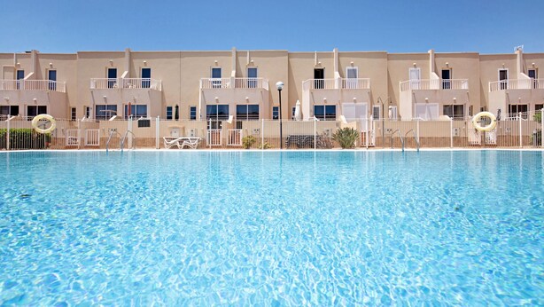 A swimming pool with clear blue water, surrounded by a fence, and a row of beige multi-story buildings with balconies.