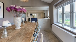 A dining area with a wooden table, chairs, a vase with flowers, and a window with a view of greenery. - Pen y Bryn -  a cottage that sleeps 10 guests  in 5 bedrooms (Pwllheli)