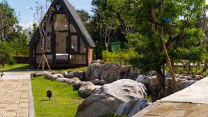 A wooden cabin with a sloped roof, surrounded by a stone pathway, large rocks, and green grass.