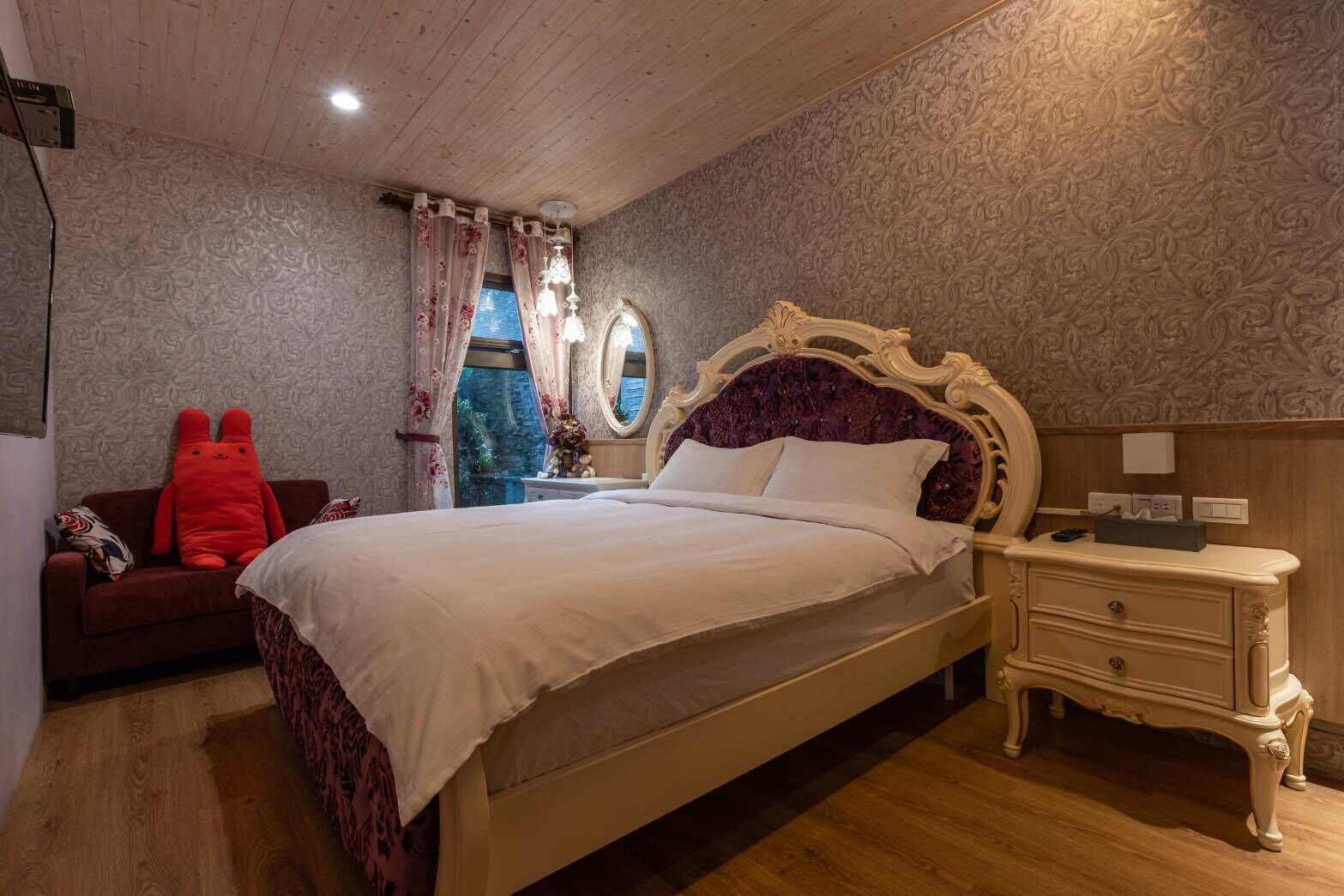 A bedroom with a large bed, a nightstand, a dresser, a red plush toy, and a window with curtains.