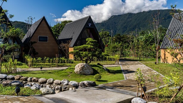 A row of wooden cabins with sloped roofs, surrounded by lush greenery and a stone pathway.
