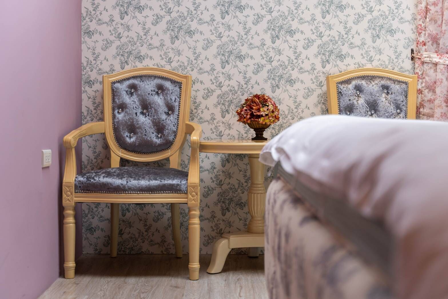 Una habitación con papel tapiz floral, una silla de madera con asiento acolchado, una mesita con un florero y una cama con almohada blanca.