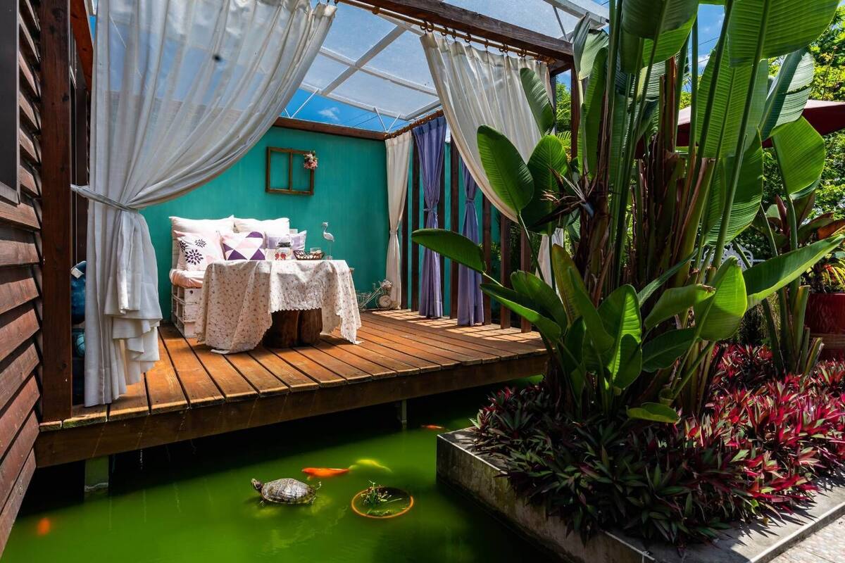 Un área de estar acogedora al aire libre con una terraza de madera, una mesa con mantel blanco y una silla. Hay una pequeña tortuga en el suelo y un ave en el agua.