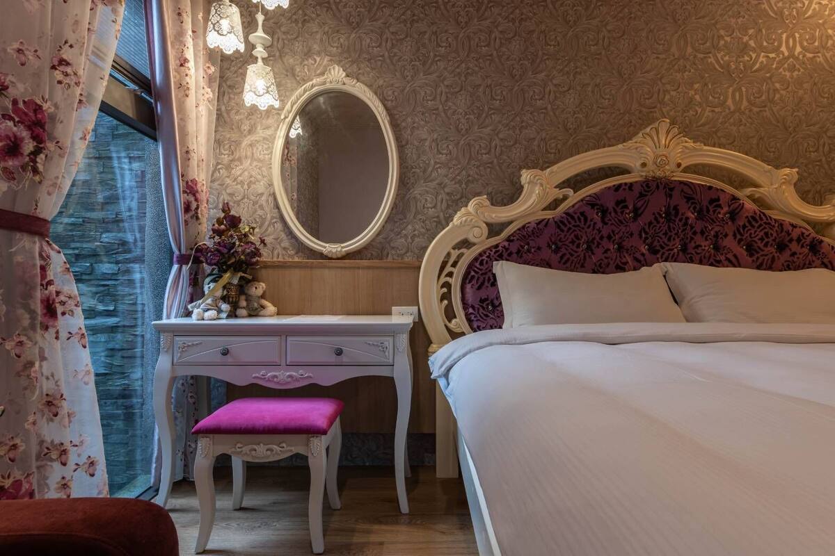 Un dormitorio con una cama, un tocador con espejo, una silla rosa y un aparador.