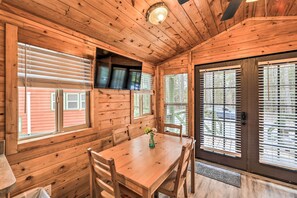 L’intérieur d’un chalet en bois comprend une table à manger, des chaises, un téléviseur fixé au mur et une porte avec des stores.