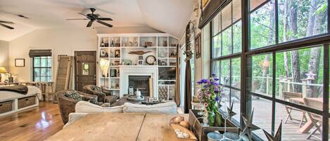 A spacious living room with a wooden table, comfortable seating, a fireplace, and large windows offering a view of the outdoors.