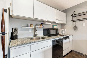 A compact kitchen with white cabinets, a granite countertop, a built-in dishwasher, a microwave, and a coffee maker. - River View Condo- Edge of Downtown (Bend)