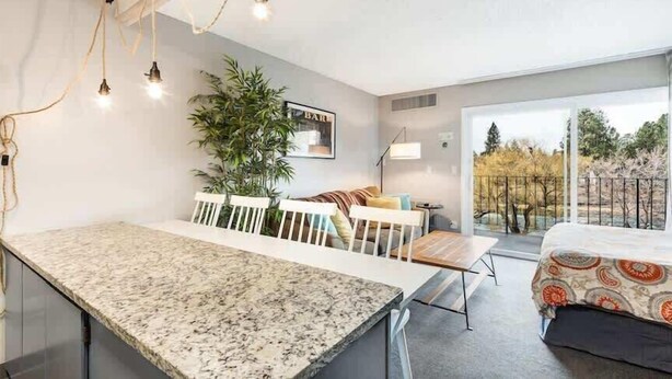 A modern kitchen with a marble countertop, a dining area with white chairs, a large potted plant, and a balcony with a view. - River View Condo- Edge of Downtown (Bend)