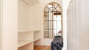 A room with a wooden floor, a grey tufted bench, and a white wardrobe with open shelves. - SKY DE LOIOS - Charming apartment with balcony (Porto)
