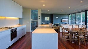 A modern kitchen with a central island, built-in oven, and a dining area with wooden chairs and a table. -  Emanjay: Walk to Beach, Million Dollar Bay Views! Mineral Spa (Mount Martha)