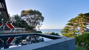 A modern outdoor pool area with a clear view of the sea, surrounded by lush greenery and red lounge chairs. -  Emanjay: Walk to Beach, Million Dollar Bay Views! Mineral Spa (Mount Martha)