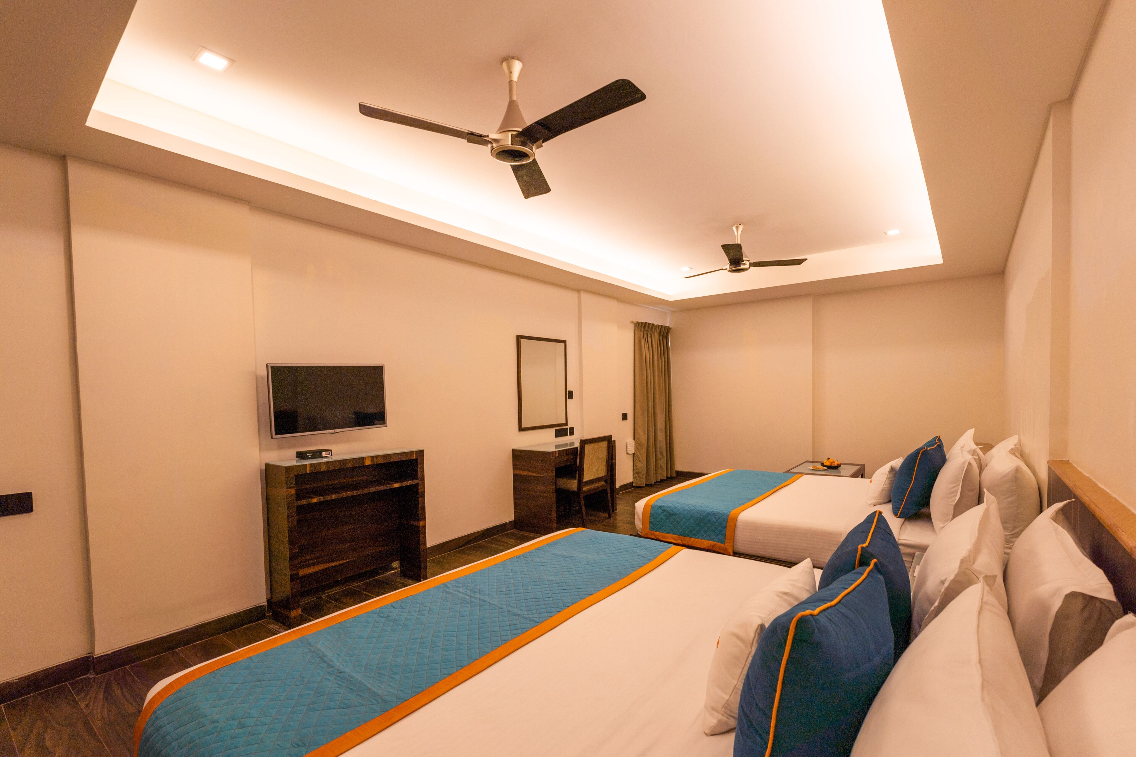 Habitación de hotel con dos camas, televisor, escritorio y ventiladores de techo.