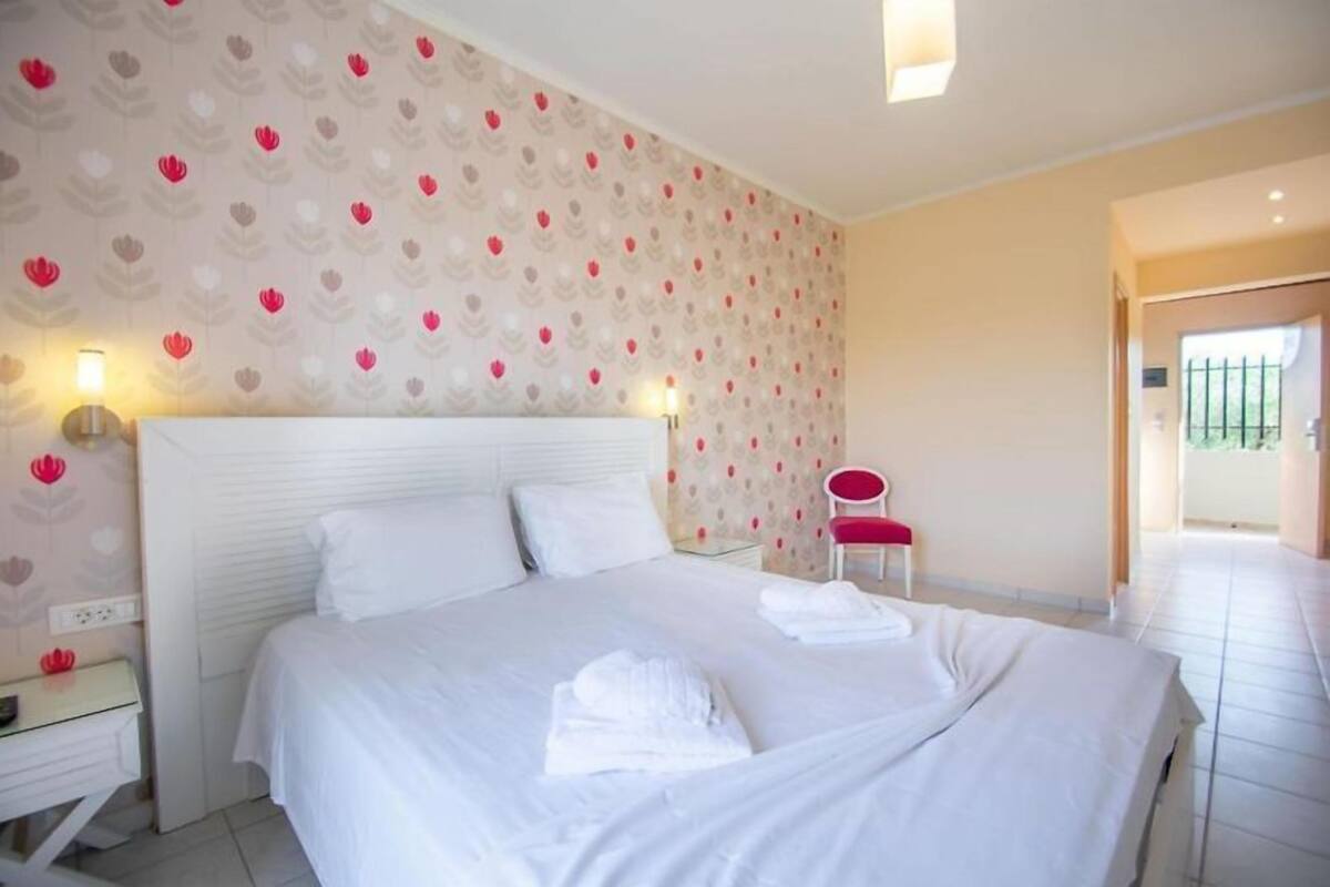 A neatly made bed with white linens and towels, a bedside table with a lamp, and a chair with a red cushion.