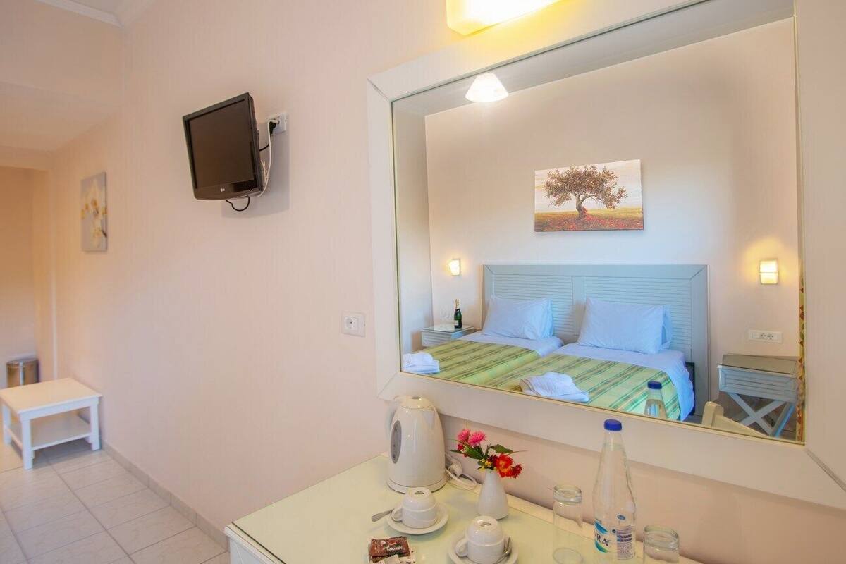 A hotel room with a bed, a bedside table with a kettle, a vase with flowers, and a large mirror reflecting the room.