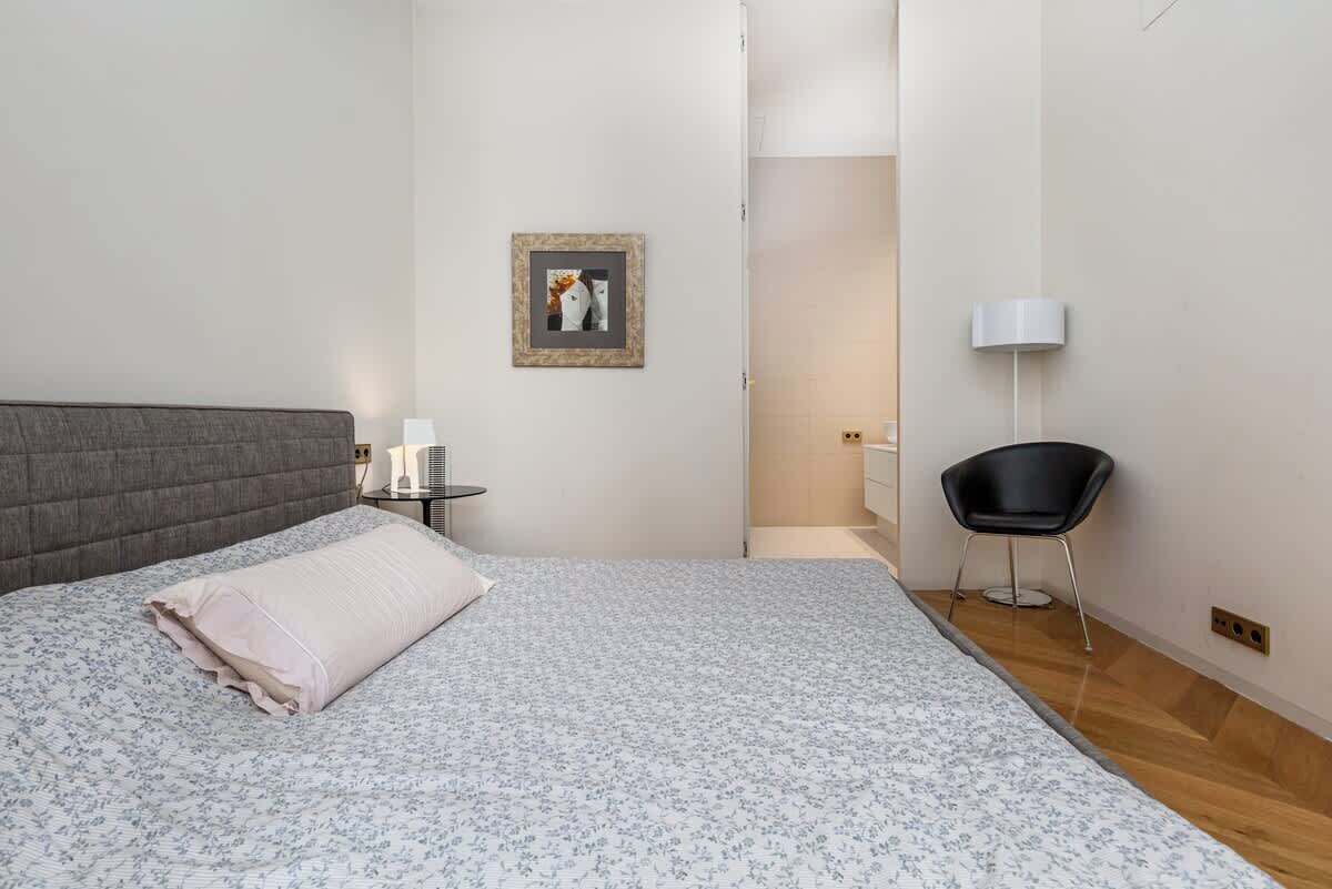 A bedroom with a bed, a bedside table, a chair, a wall-mounted lamp, and a framed picture.