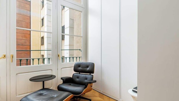 Una habitación moderna con un sofá y reposapiés de cuero negro, una pequeña mesa redonda y ventanas amplias con vistas a edificios.
