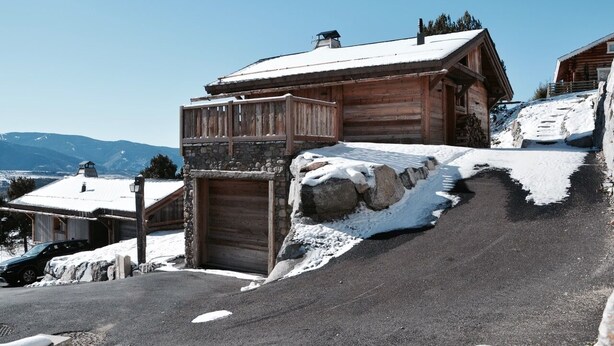 Une cabane en bois avec une entrée en pierre, un toit enneigé et une vue sur les montagnes en arrière-plan.