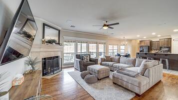 A spacious living room with a fireplace, large sofa, and ottomans. A kitchen with modern appliances is visible in the background.