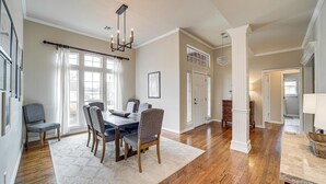 A dining area with a wooden table, chairs, and a chandelier. There are windows with curtains, a white door, and a hallway leading to another room. - Pet-Friendly Tulsa Escape w/ Private Hot Tub! (Tulsa)
