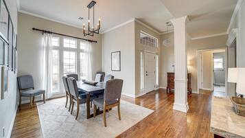 A dining area with a wooden table, chairs, and a chandelier. There are windows with curtains, a white door, and a hallway leading to another room.