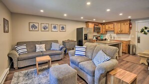 A living room with a grey sofa set, a wooden coffee table, and a kitchen area with wooden cabinets and a refrigerator. - Gilford Escape w/ Boat Slip on Sanders Bay! (Gilford)