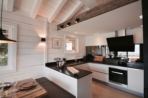 A modern kitchen with white cabinetry, a black countertop, and a large flat-screen TV. - Chalet Opale Collection Perles (BOLQUÈRE FONT-ROMEU)