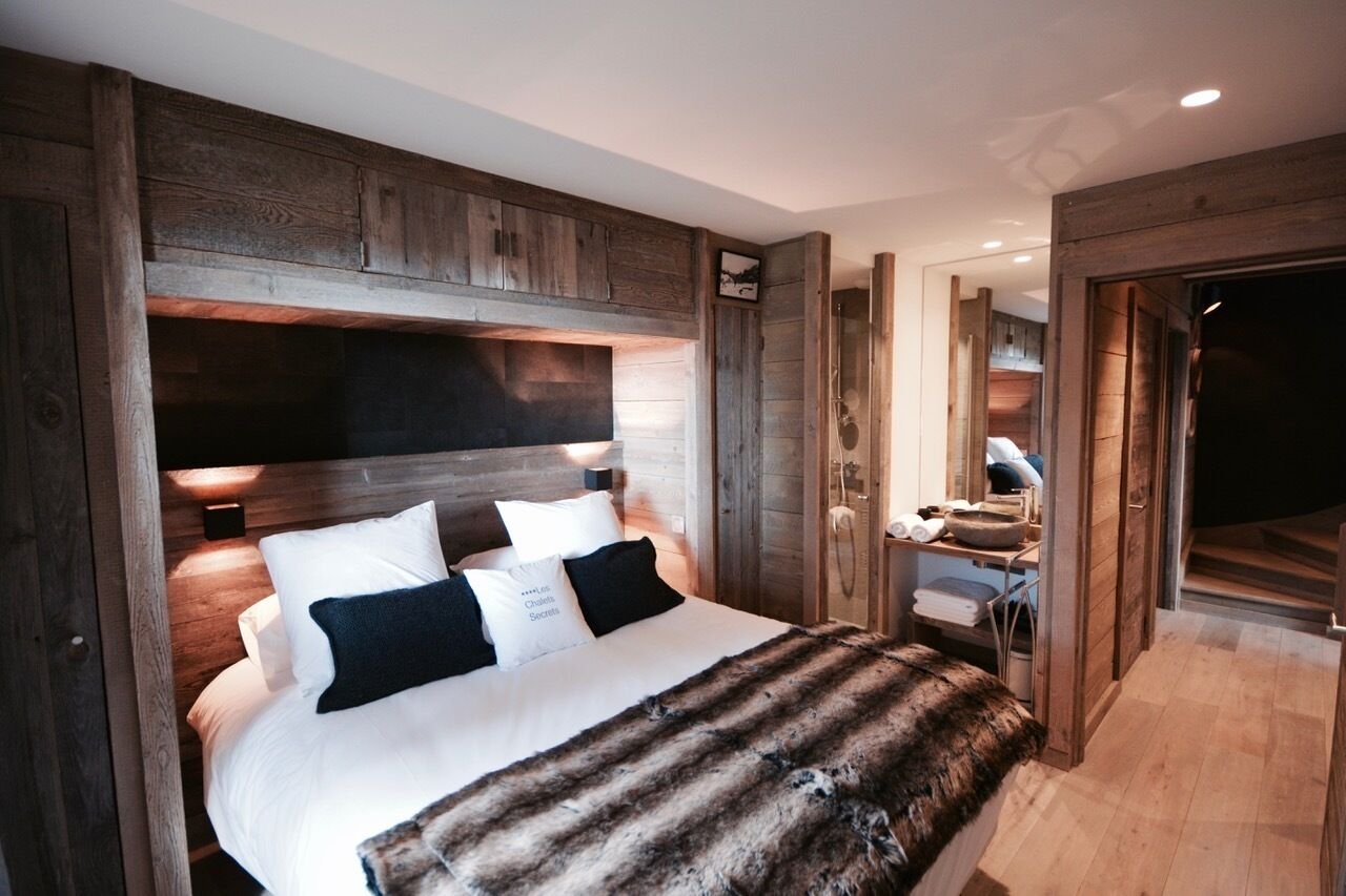 Une chambre avec un grand lit, une tête de lit en bois et un plaid en fourrure. On y trouve un bureau avec une chaise et un miroir.