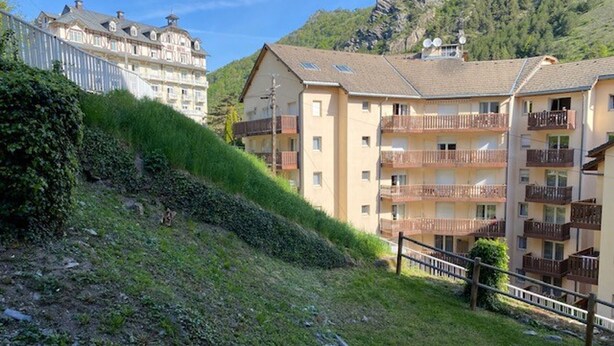 Una zona residencial en una ladera con edificios de varios pisos, algunos con balcones y antenas parabólicas, y una pendiente cubierta de césped con una valla de madera.