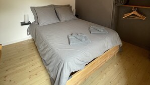Een slaapkamer met een houten plafond, een bed met grijze beddengoed, een nachtkastje met een lamp, en een wandplank met een handdoek en ophanger.