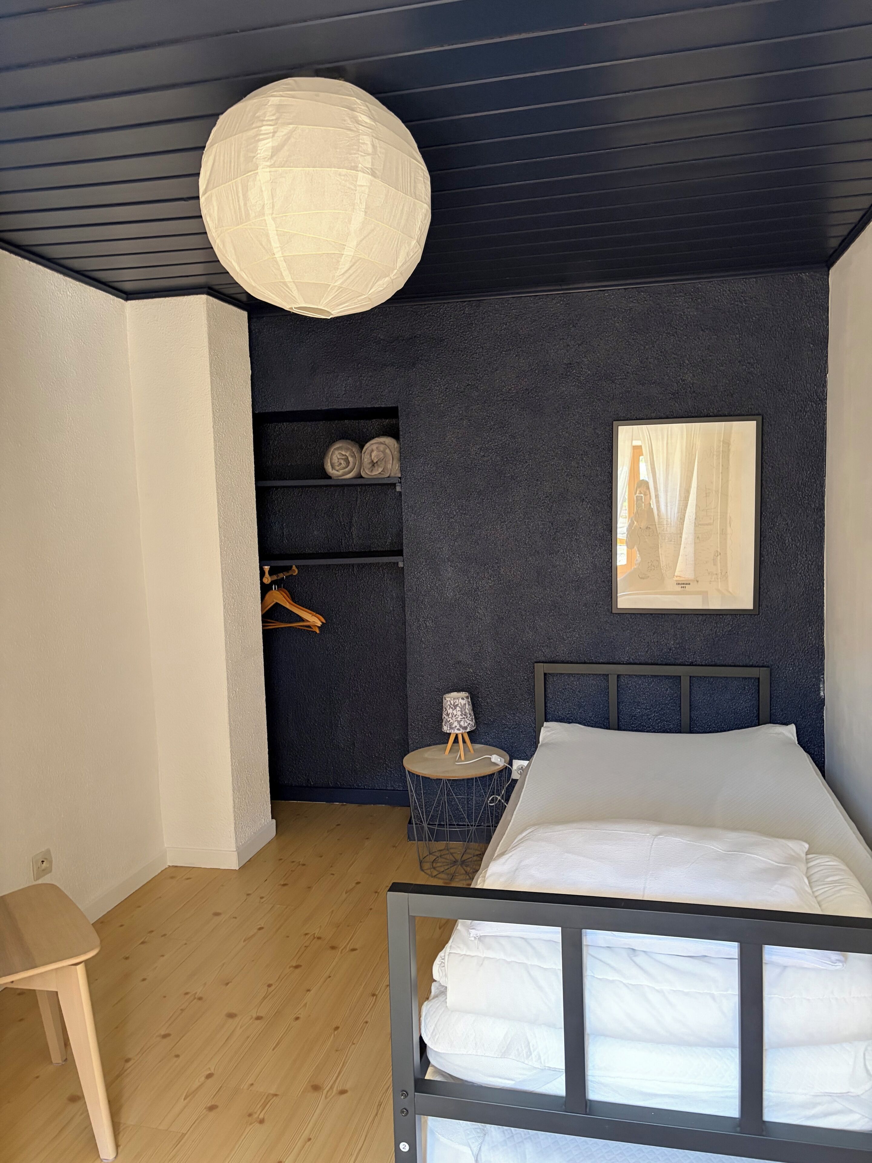 Un dormitorio con cama, mesita de noche, lámpara colgante, cuadro enmarco y armario con estantes.