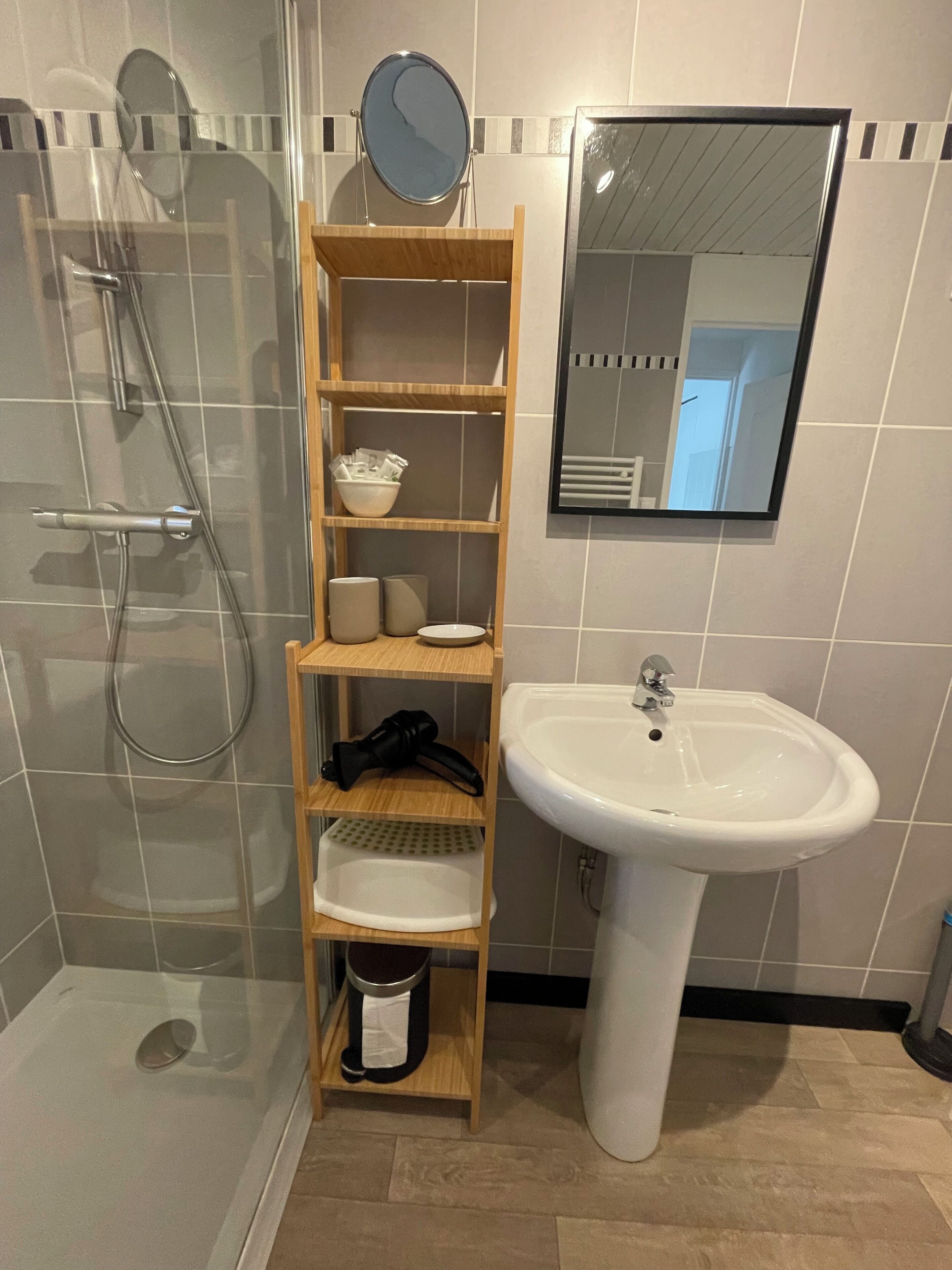 Une salle de bain avec une douche à parois de verre, une étagère en bois où sont rangés les articles de toilette, un lavabo blanc et un miroir.