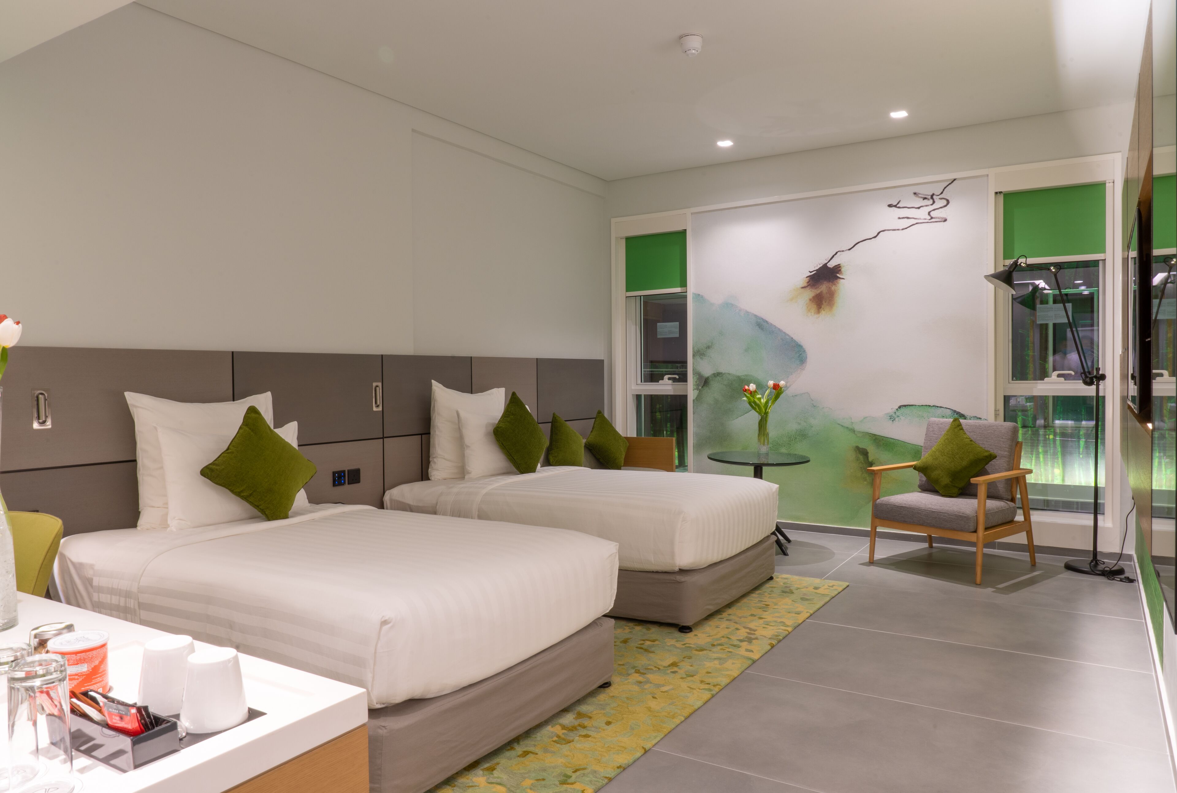 A modern hotel room with two beds, a small table, a chair, and a view of the outdoors.