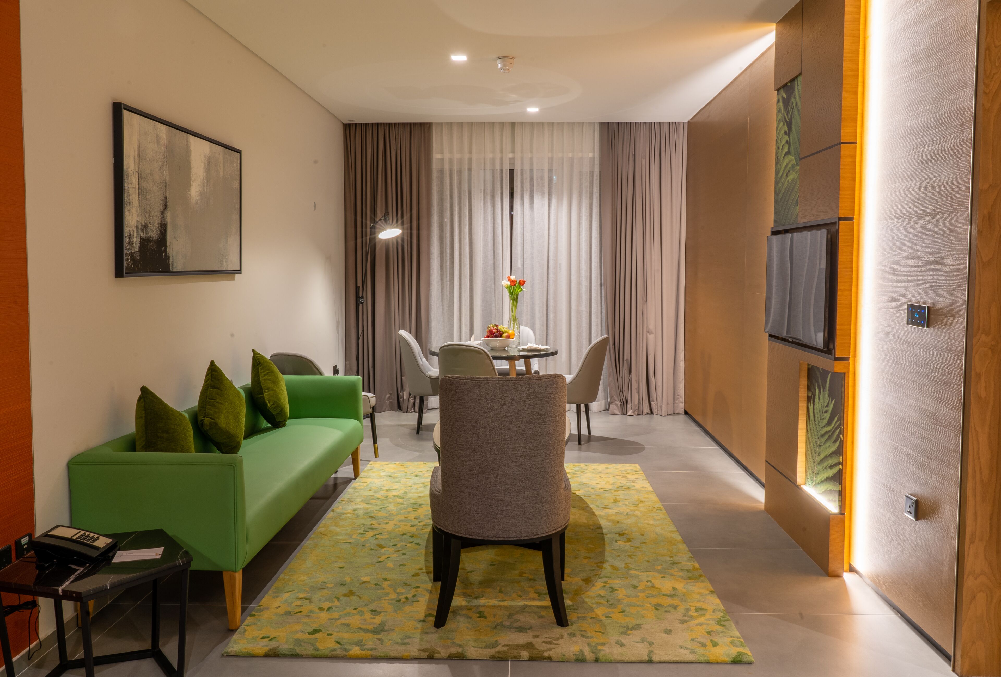 Een moderne hotelkamer met een groene bank, een eettafel met stoelen, een klein bijzettafeltje en een groot raam met gordijnen.
