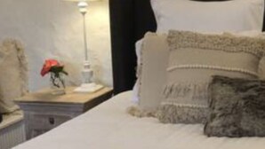 Een keurig opgemaakt bed met witte lakens en meerdere kussens, een bedlampje en een klein decoratief object op het nachtkastje.