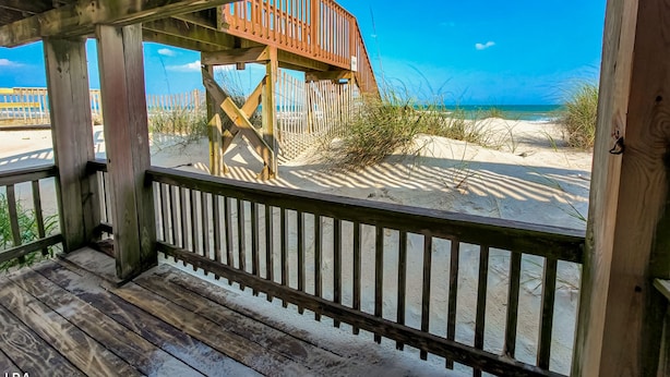 Une terrasse en bois dotée d’une rambarde, qui offre une vue sur une plage de sable, et un escalier en bois menant à une plateforme plus élevée.