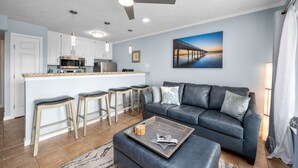 Una sala moderna con un sofá de cuero oscuro, taburetes de madera, una mesa de centro y una cocina con armarios blancos y un microondas.