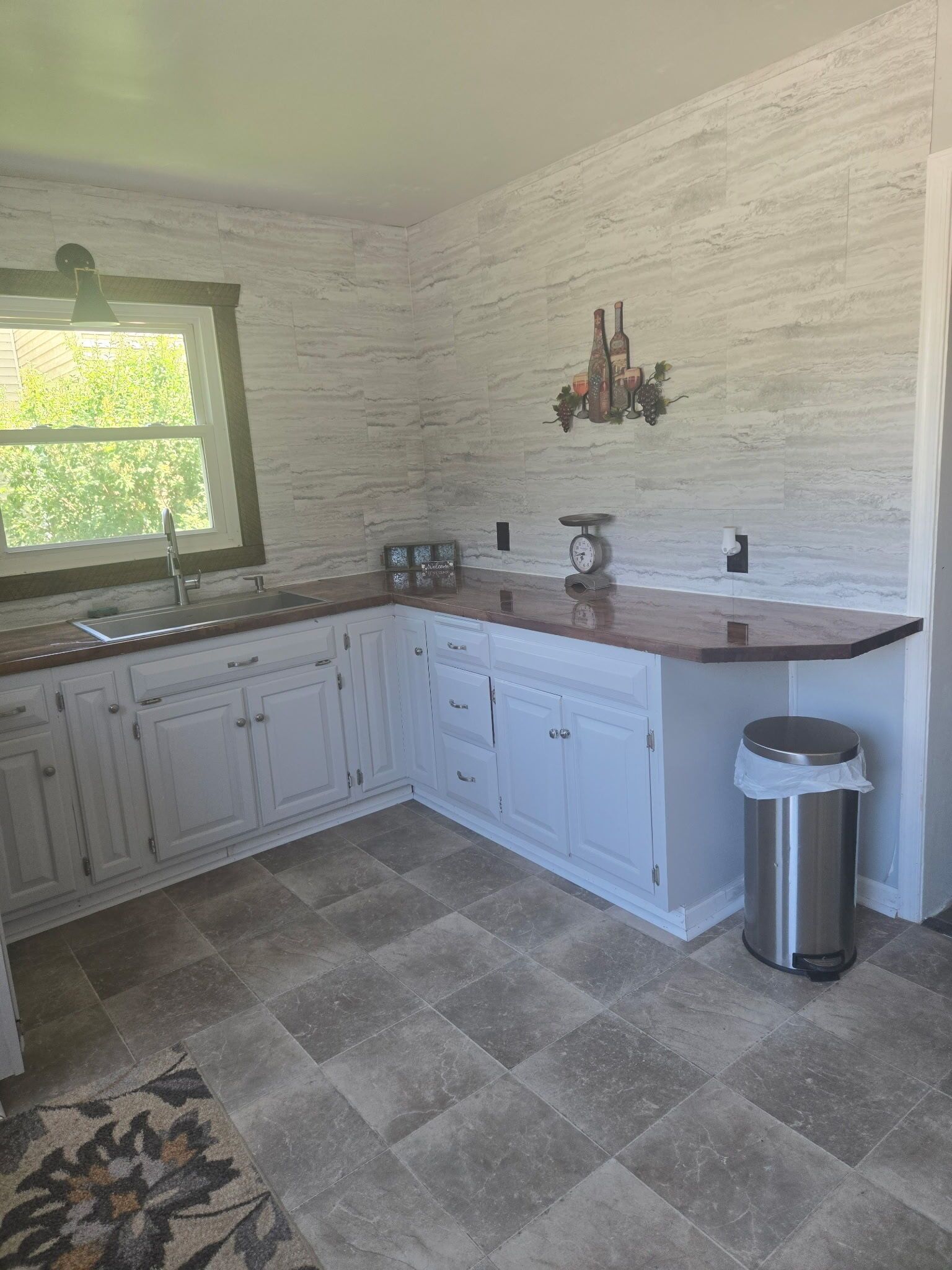 A kitchen with white cabinets, a stainless steel sink, a clock, and a trash can.