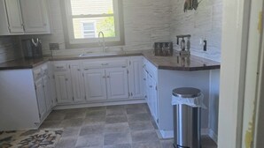 Una cucina con mobili bianchi, una finestra, un cestino in acciaio inossidabile e un pavimento piastrellato.