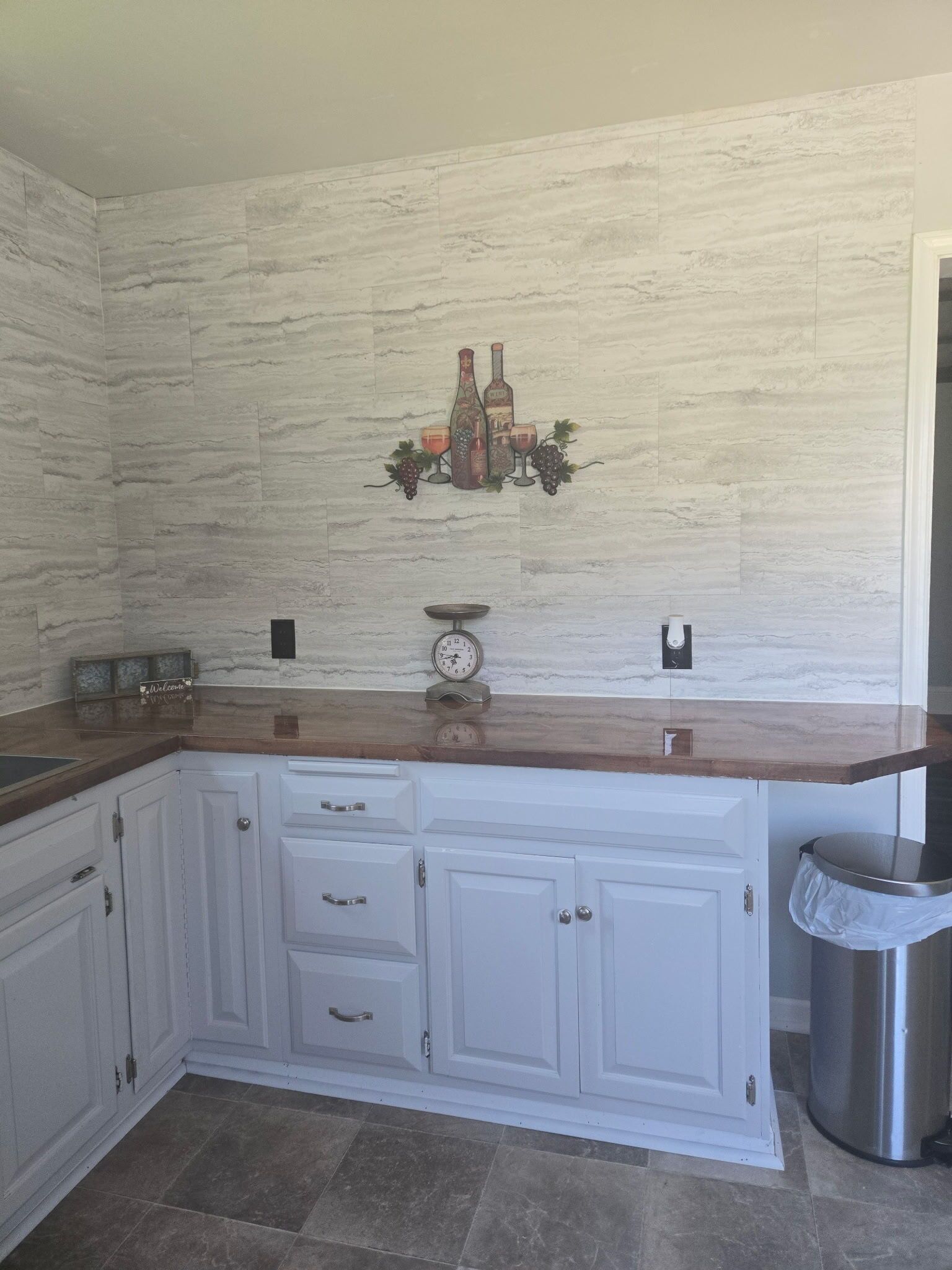 A kitchen with white cabinets, a wooden countertop, a wall clock, and a wine-themed decoration.