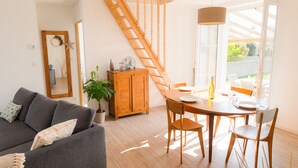 Uma sala de estar moderna com um sofá cinzento, mesa de jantar de madeira e cadeiras. Uma escada de madeira leva a uma área externa.