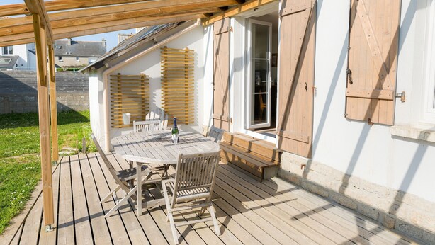 Um deck de madeira com mesa e cadeiras, uma pérgula e vista para uma rua residencial.