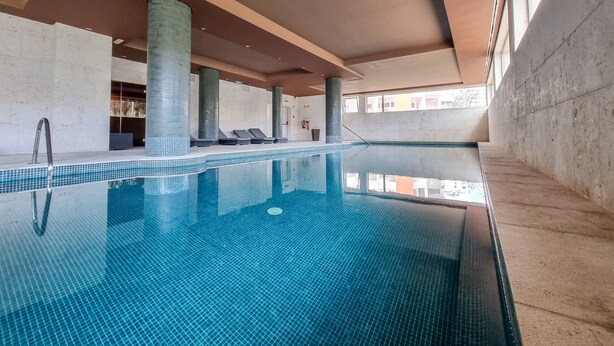 Ein Innenpool mit klarem, blauem Wasser, gefliestem Boden und Liegestühlen am Rand.