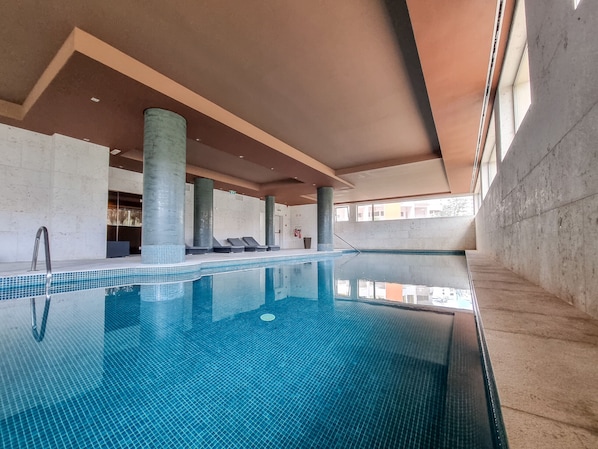 An indoor swimming pool with clear blue water, tiled floor, and lounge chairs along the edge.