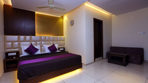 A modern bedroom with a large bed, a ceiling fan, a wall clock, and a small seating area. - Hotel Hindustan Residency (Thane)